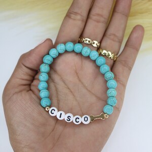 Puede incluir: Pulsera de cuentas turquesas con detalles dorados y un dije de hueso. La pulsera tiene cuentas de letras blancas que deletrean el nombre "CISCO".