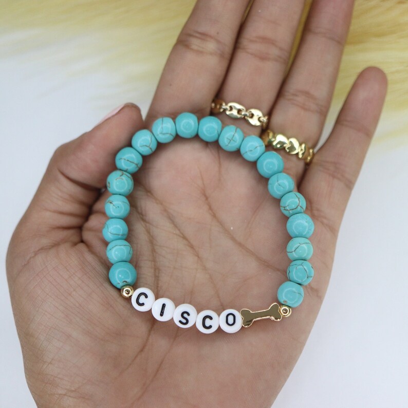 Puede incluir: Pulsera de cuentas turquesas con detalles dorados y un dije de hueso. La pulsera tiene cuentas de letras blancas que deletrean el nombre "CISCO".