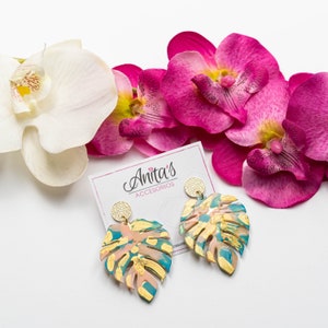 Puede incluir: Un par de pendientes dorados y turquesa con un dise&ntilde;o de hoja tropical. Los pendientes est&aacute;n sobre una superficie blanca con orqu&iacute;deas rosas.