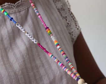 Puede incluir: Un cordón de cuentas de colores con el nombre "MISS KISER" escrito en letras blancas. El cordón tiene una cuenta de madera en el extremo y un cierre metálico.