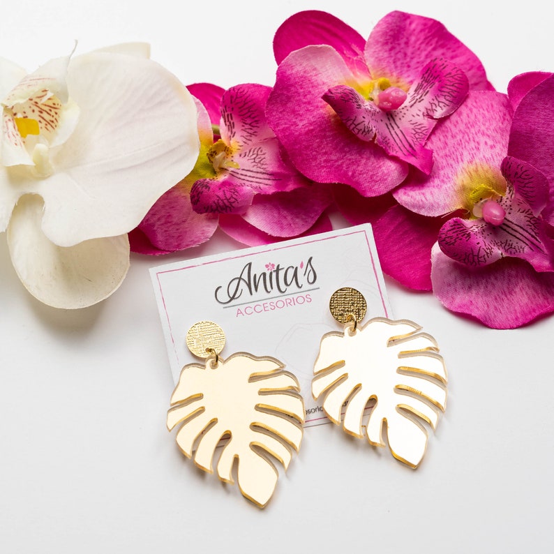 Puede incluir: Pendientes de tono dorado con dise&ntilde;o de hoja de monstera. Los pendientes est&aacute;n sobre una superficie blanca con flores de orqu&iacute;dea rosa.