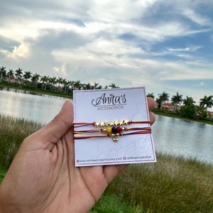 May include: A set of three friendship bracelets with red, yellow, and blue string, gold star beads, and a small heart charm. The bracelets are displayed on a white card with the text "Anita's Accesorios" and website information.