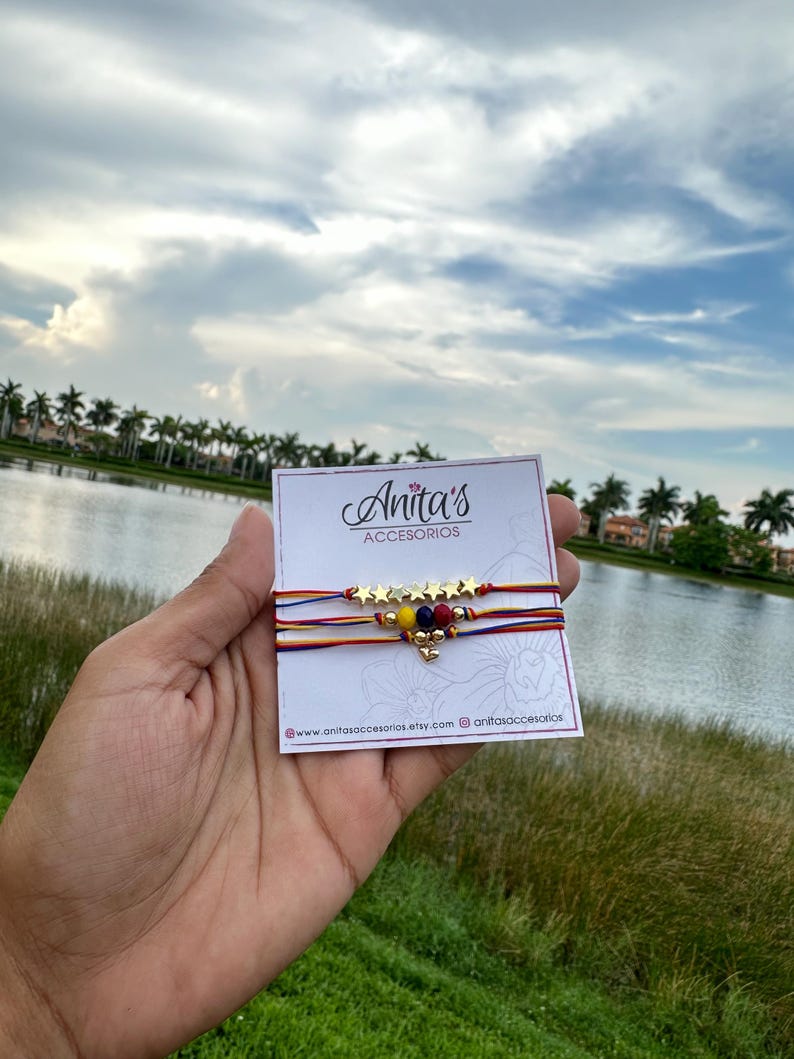 May include: A set of three friendship bracelets with red, yellow, and blue string, gold star beads, and a small heart charm. The bracelets are displayed on a white card with the text "Anita's Accesorios" and website information.