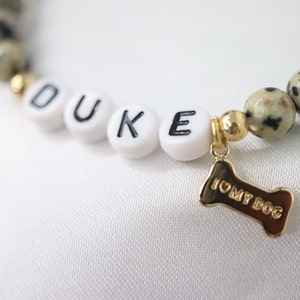 Peut inclure: Un bracelet de perles avec le nom "DUKE" &eacute;pel&eacute; en perles blanches. Le bracelet comprend des accents dor&eacute;s et un charm en forme d'os dor&eacute; avec l'inscription "I ❤️ MY DOG". Les perles sont un m&eacute;lange de blanc et de beige tachet&eacute;.