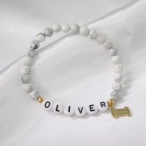 Peut inclure: Bracelet de perles blanches et grises avec le nom "OLIVER" &eacute;pel&eacute; en perles de lettres blanches. Un charm en forme d'os de couleur dor&eacute;e est suspendu au bracelet. Le bracelet est pos&eacute; sur une surface en satin blanc.
