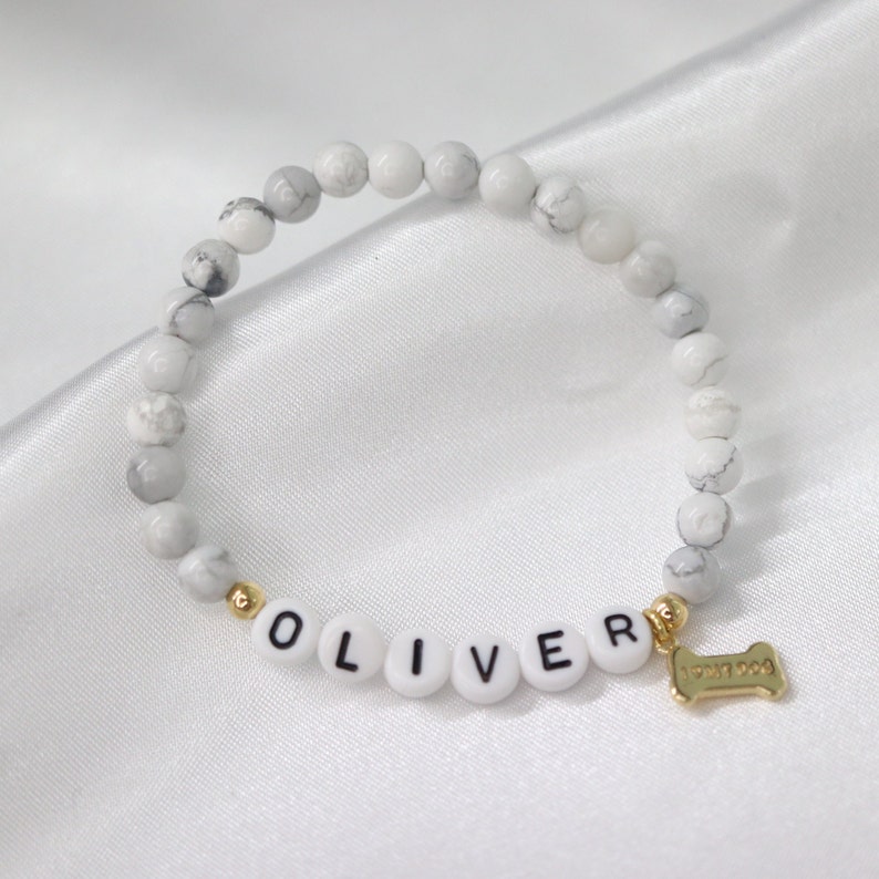 Peut inclure: Bracelet de perles blanches et grises avec le nom "OLIVER" &eacute;pel&eacute; en perles de lettres blanches. Un charm en forme d'os de couleur dor&eacute;e est suspendu au bracelet. Le bracelet est pos&eacute; sur une surface en satin blanc.