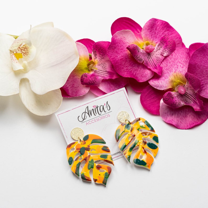 Puede incluir: Un par de pendientes de hoja de monstera amarilla, verde y naranja con detalles dorados. Los pendientes se muestran sobre una superficie blanca con flores de orqu&iacute;dea rosa y una tarjeta que dice "Anita's Acessorios".