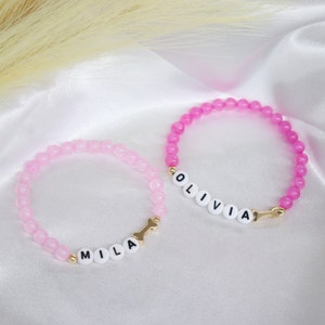 Puede incluir: Dos pulseras de cuentas rosadas con dijes dorados y cuentas de letras blancas que deletrean los nombres "Olivia" y "Mila".