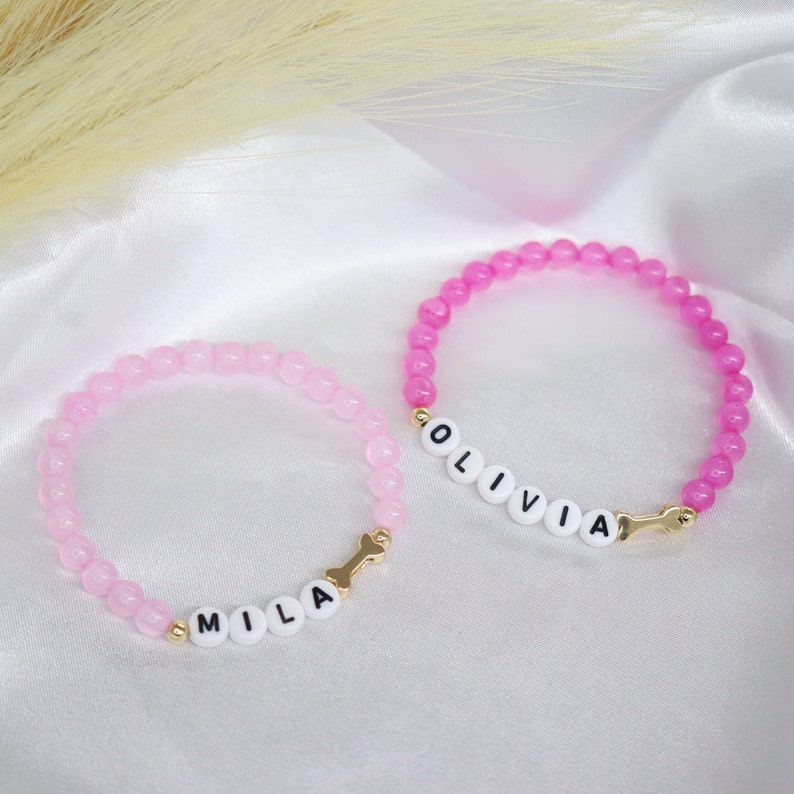 Puede incluir: Dos pulseras de cuentas rosadas con dijes dorados y cuentas de letras blancas que deletrean los nombres "Olivia" y "Mila".