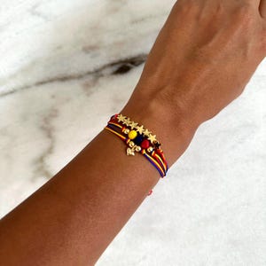 May include: A colorful string bracelet with gold stars and beads. The bracelet is red, yellow, and blue, and has a small gold charm.