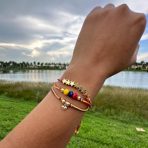 May include: Three bracelets with red, yellow, and blue string, gold star charms, and gold beads. The bracelets have a small gold heart charm.