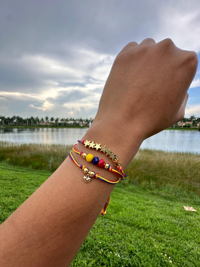 May include: Three bracelets with red, yellow, and blue string, gold star charms, and gold beads. The bracelets have a small gold heart charm.
