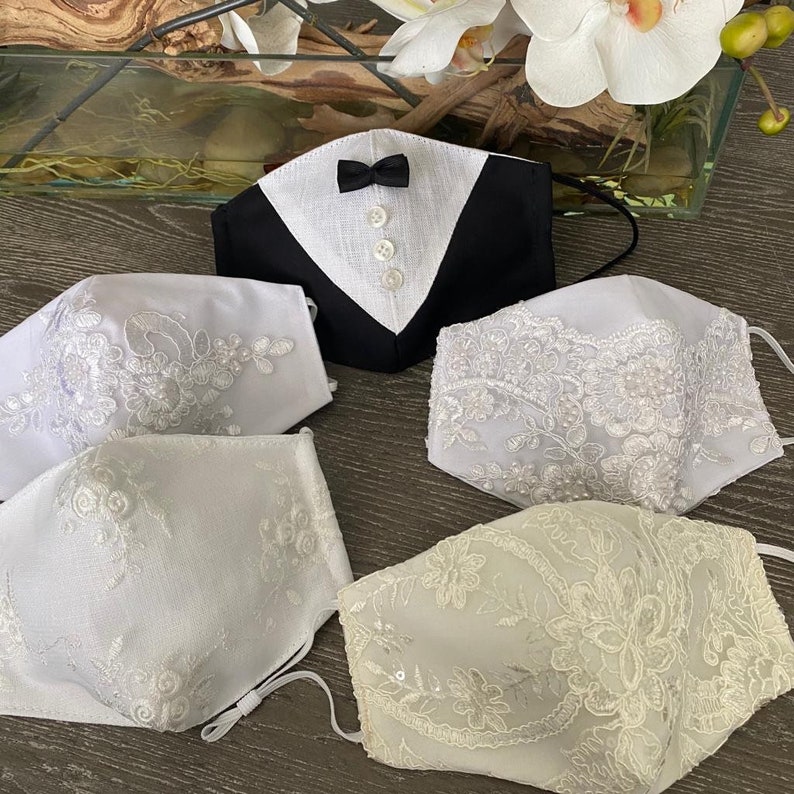 May include: Four white face masks with lace detailing. One mask features a black tuxedo design with a white bow tie.