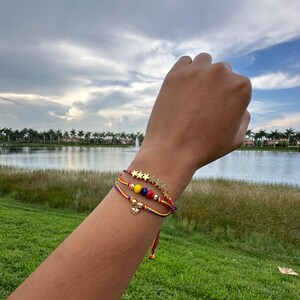 May include: Three colorful string bracelets with gold stars and beads. The bracelets are worn on a person's wrist.