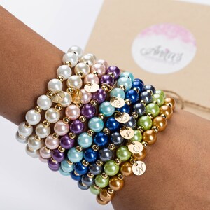 May include: A stack of colorful pearl bracelets with gold charms, each charm has a letter engraved on it. The bracelets are on a person's wrist.