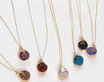 Könnte beinhalten: Sechs goldene Halsketten mit verschiedenen Edelsteinanhängern in unterschiedlichen Farben. Jeder Anhänger hat einen goldenen Buchstaben-Anhänger. Die Farben der Edelsteine sind lila, schwarz, weiß, rot, blau und hellbraun.