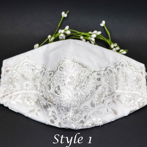 May include: White face mask with a white lace trim and pearl embellishments. The mask is on a black background. Style 1.