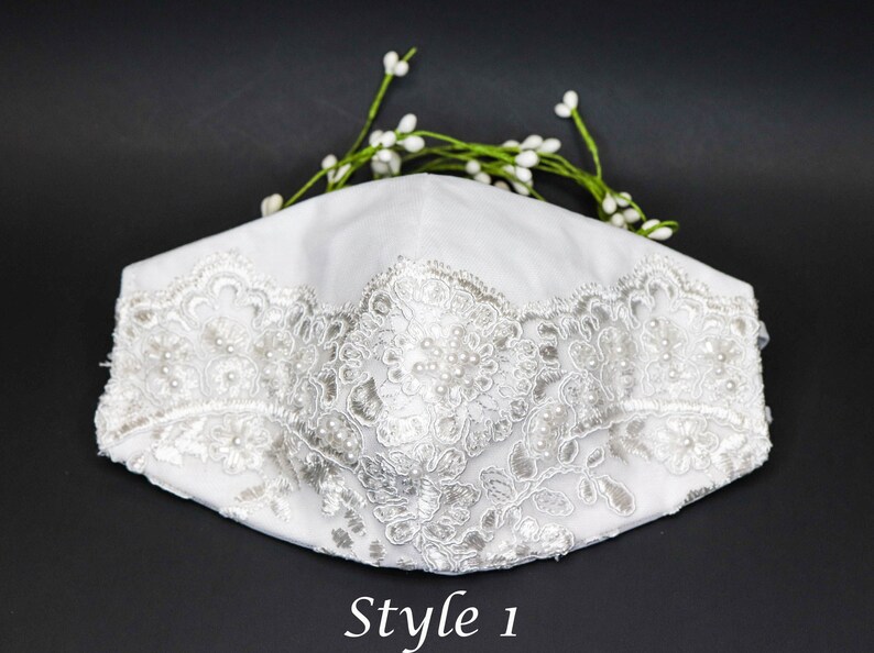 May include: White face mask with a white lace trim and pearl embellishments. The mask is on a black background. Style 1.