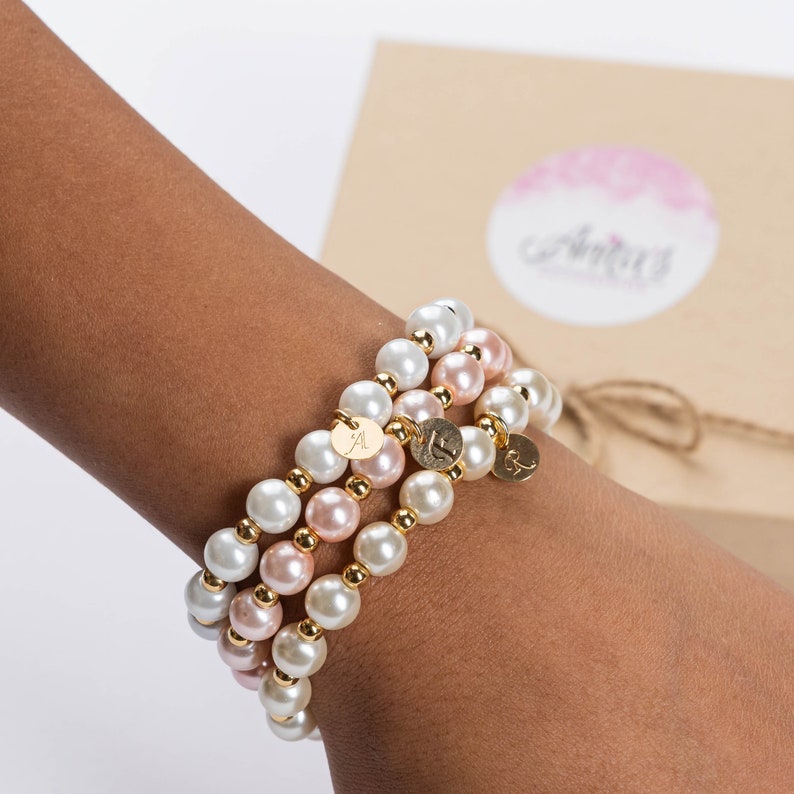 May include: Three layered bracelets with white and pink pearls and gold accents. Each bracelet has a small gold charm with a letter on it.