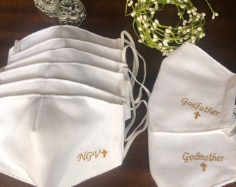 May include: A stack of six white fabric face masks with gold embroidery. The top mask says "NGV+" and the bottom mask says "Godmother" with a small cross. The masks have elastic ear loops.