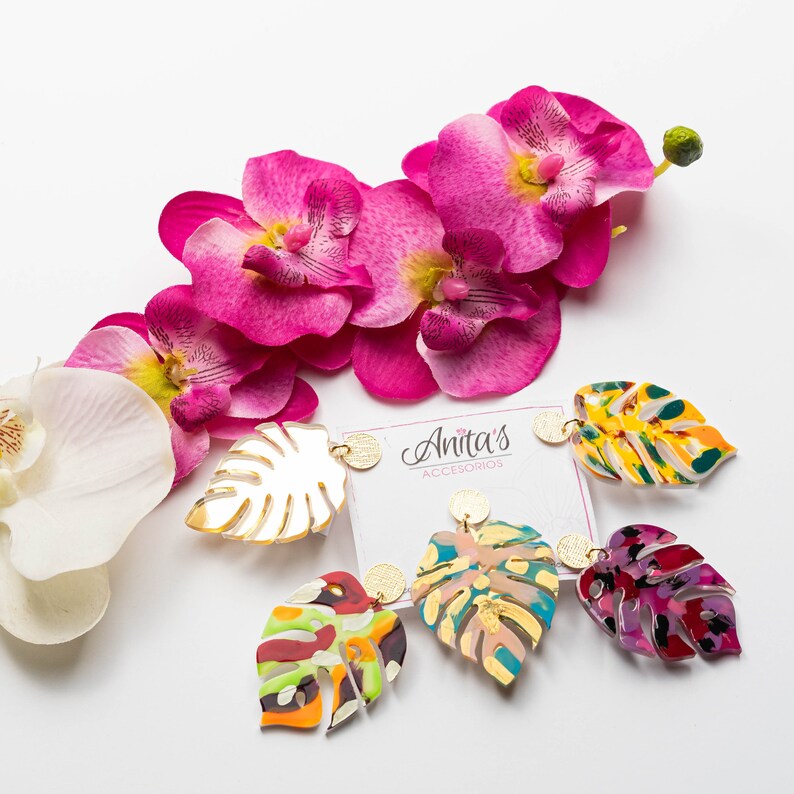 Puede incluir: Cuatro pares de pendientes de hojas tropicales de colores con detalles dorados. Los pendientes se muestran sobre una superficie blanca con flores de orqu&iacute;dea rosa.