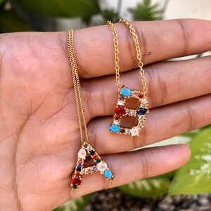 Puede incluir: Dos collares de oro con colgantes de letras de piedras preciosas de colores. Los collares presentan las letras A y B, cada una adornada con una variedad de piedras preciosas de colores.