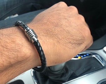 Puede incluir: Un brazalete de cuero negro con un cierre magnético plateado. El brazalete está trenzado y tiene un diseño elegante y moderno.