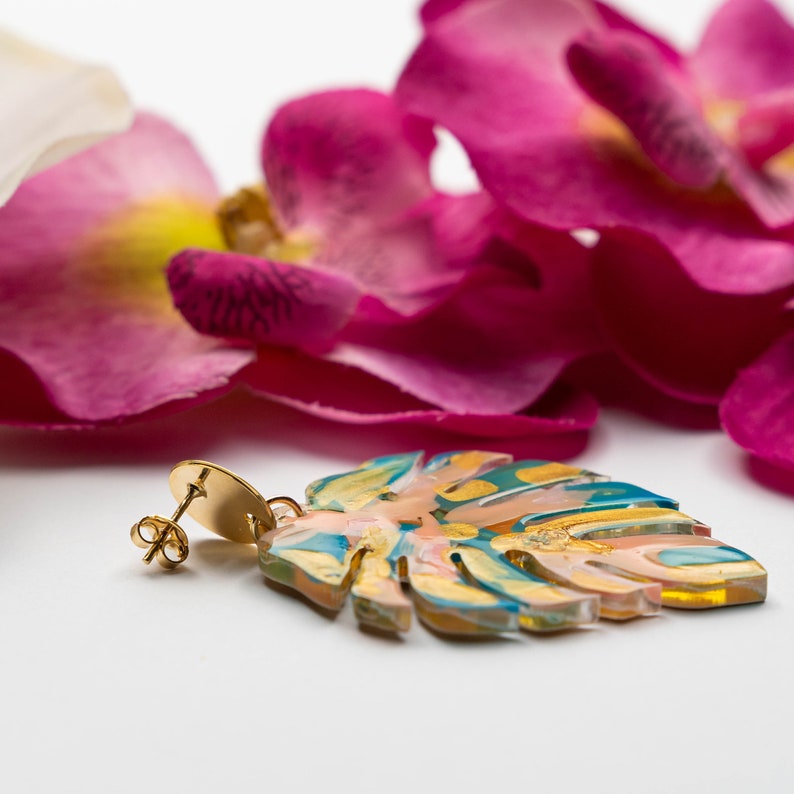 Puede incluir: Pendiente de tono dorado con un dise&ntilde;o de hoja transl&uacute;cida y colorida. La hoja es de color rosa claro, azul y amarillo con detalles dorados. El pendiente est&aacute; sobre un fondo blanco.