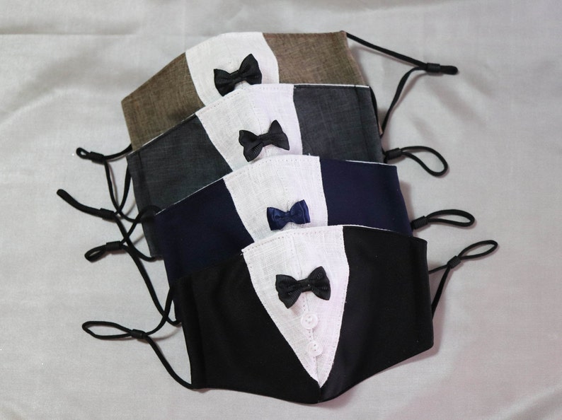 May include: Five face masks with different colored fabrics and bow ties. The masks are layered on top of each other, with the black mask on the bottom and the brown mask on top. The bow ties are all different colors, including black, navy blue, and white.