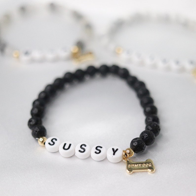 Peut inclure: Bracelet de perles de lave noires avec des perles de lettres blanches &eacute;pelant "SUSSY". Un charm dor&eacute; en forme d'os de chien est attach&eacute;. D'autres bracelets sont visibles en arri&egrave;re-plan.