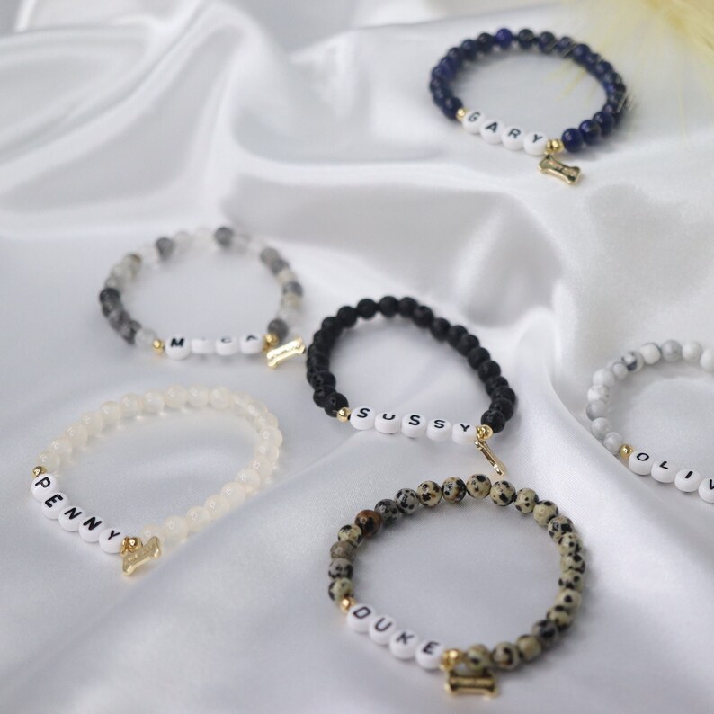 Peut inclure: Plusieurs bracelets de perles avec des lettres blanches &eacute;pelant des noms tels que "Penny", "Duke" et "Gary". Chaque bracelet a un charme dor&eacute;, certains en forme d'os. Les bracelets sont de diff&eacute;rentes couleurs, dont le noir, le blanc et le mouchet&eacute;.
