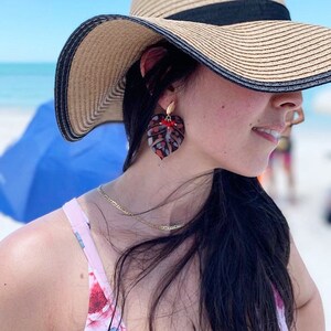 Puede incluir: Una mujer con un sombrero de paja y un traje de ba&ntilde;o de estampado floral con grandes pendientes en forma de hoja roja y negra.