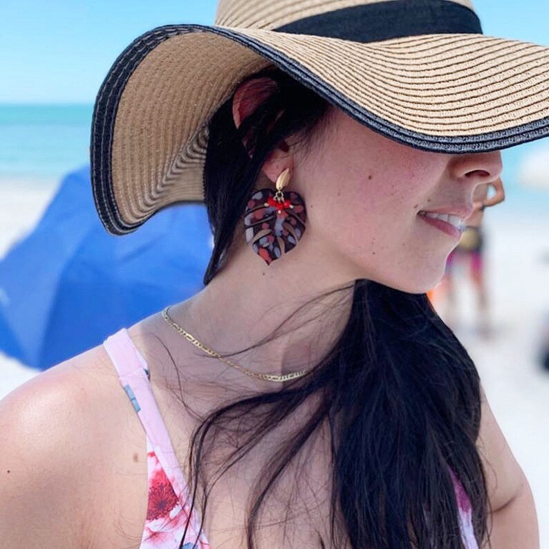 Puede incluir: Una mujer con un sombrero de paja y un traje de ba&ntilde;o de estampado floral con grandes pendientes en forma de hoja roja y negra.
