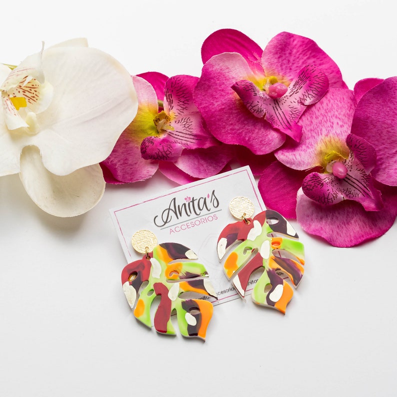 Puede incluir: Un par de pendientes de hoja de monstera coloridos con detalles dorados. Los pendientes est&aacute;n hechos de arcilla polim&eacute;rica y tienen un esquema de color verde, rojo, naranja y blanco. Los pendientes cuelgan de un fondo blanco con flores de orqu&iacute;dea rosa.