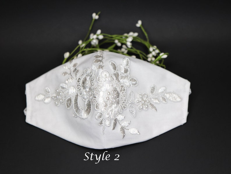 May include: White face mask with delicate white lace embroidery and pearl accents. The mask has a simple design and is perfect for a wedding or special occasion.