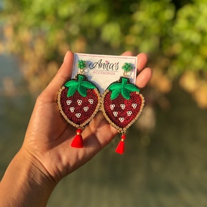 May include: A pair of red and green strawberry-shaped earrings with gold accents and red tassels. The earrings are made of fabric and sequins. The earrings are on a white card with the text "Anita's Accesorios".