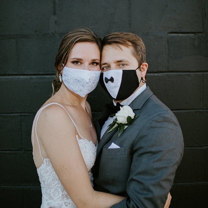 May include: A bride and groom wearing white and black face masks with a white flower on the groom's lapel. The couple is standing in front of a black brick wall.