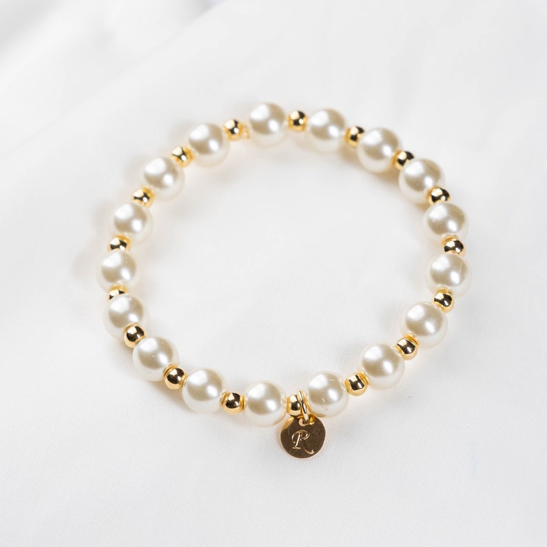 May include: A white pearl bracelet with gold beads and a gold charm with the letter 'R' on it.