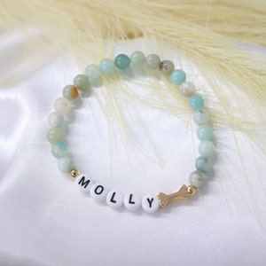 Puede incluir: Un brazalete de cuentas verdes y blancas con el nombre "MOLLY" escrito en letras blancas y un dije de hueso dorado.