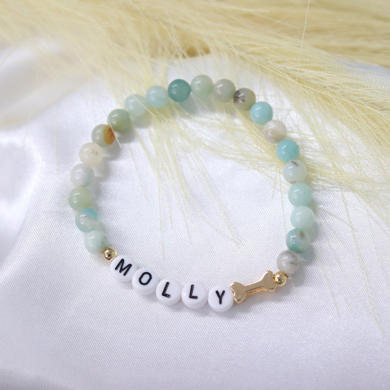 Puede incluir: Un brazalete de cuentas verdes y blancas con el nombre "MOLLY" escrito en letras blancas y un dije de hueso dorado.
