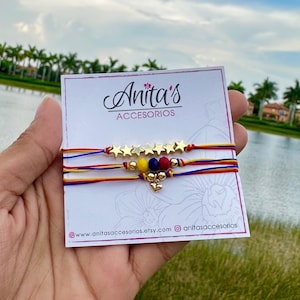 May include: Three colorful string bracelets with gold star charms and a small gold heart charm. The bracelets are displayed on a white card with the text "Anita's Accesorios" and website information.