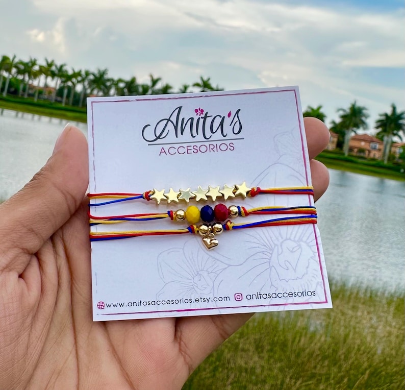 May include: Three colorful string bracelets with gold star charms and a small gold heart charm. The bracelets are displayed on a white card with the text "Anita's Accesorios" and website information.