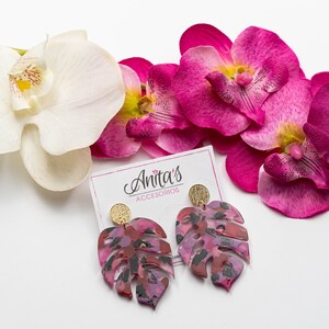 Puede incluir: Un par de pendientes de hoja de monstera rosa y negra con detalles dorados. Los pendientes est&aacute;n sobre una superficie blanca con flores de orqu&iacute;dea rosa.