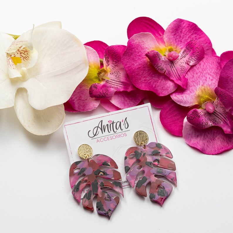 Puede incluir: Un par de pendientes de hoja de monstera rosa y negra con detalles dorados. Los pendientes est&aacute;n sobre una superficie blanca con flores de orqu&iacute;dea rosa.