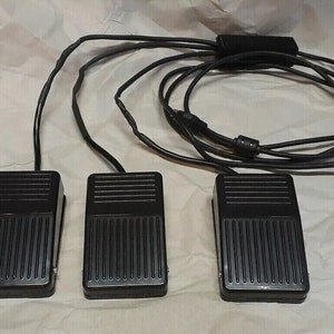 Puede incluir: Tres pedales negros con superficies estriadas, conectados por cables negros y un cable central. Los pedales son rectangulares y parecen estar hechos de plástico o un material similar. Los cables están agrupados.