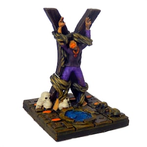 Puede incluir: Una figura en miniatura de una persona atada y amarrada a una cruz de madera, con cuerda y cráneos que rodean la base. La figura lleva una túnica morada y tiene el pelo naranja. La base es una piedra marrón oscura con un charco de agua azul en el centro.