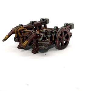May include: A miniature wooden catapult with a brown and green paint scheme. The catapult has a single wheel and two arms that are raised up.