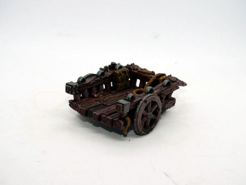 May include: A miniature wooden cart with a single wheel, painted in brown and green tones. The cart has a wooden frame and a rope tied to the back. There is a small brown bag in the cart.