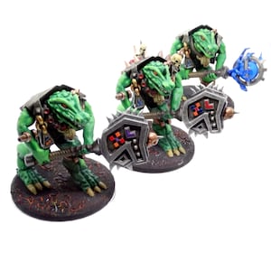 May include: Three green reptilian warrior figurines with detailed armor and weapons. Each figure has a reptilian head, holding a spiked hammer or a blue orb staff. The figures stand on textured, dark brown bases, with a white background.