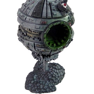 May include: A detailed model of a Death Star, a large, spherical space station from the Star Wars franchise. The model is painted in a weathered gray and green color scheme, with a large, open hatch on one side. The model is mounted on a rocky base.